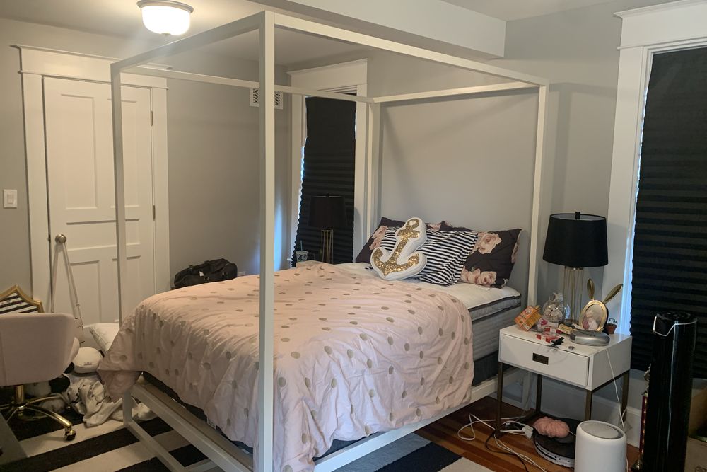 Bedroom before the design plan: white canopy bed, dark window shades, striped rug and a cluttered nightstand
